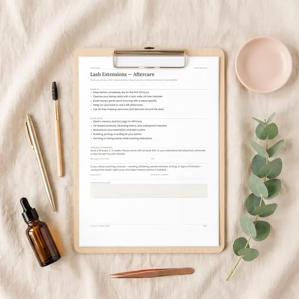 A printed Lash Extensions Aftercare sheet on a clipboard, with pens, a lash spoolie and eucalyptus on linen